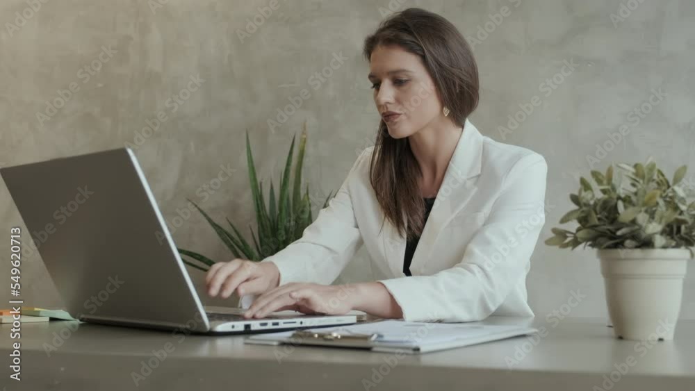 Young serious businesswoman or student in elegant white jacket making notes in copybook and looking at laptop screen while sitting by workplace in home office and teleworking Young serious businesswoman or student in elegant white jacket making notes in copybook and looking at laptop screen while sitting by workplace in home office and teleworking