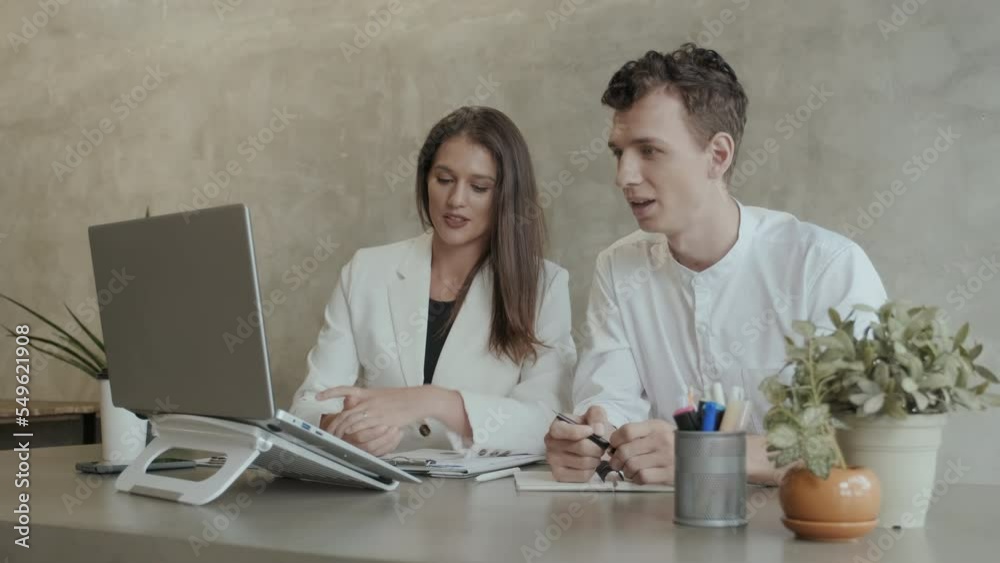 Two young employees in formalwear sitting in front of laptop during digital meeting with online colleagues while female broker making presentation of financial data