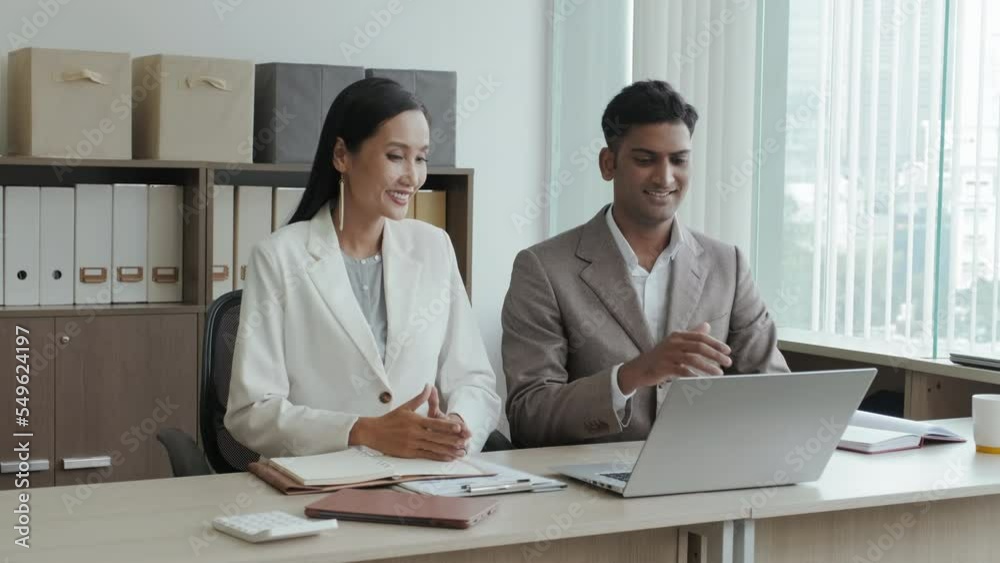 Indian businessman and his Asian female colleague sitting together at office desk, waving and talking via video call on laptop