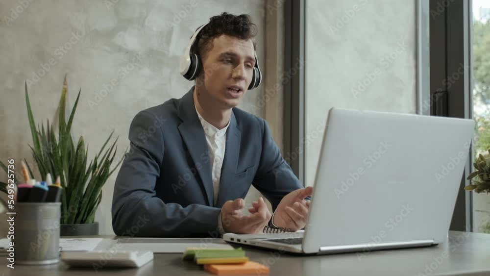 Slow motion of young modern businessman in headphones sitting by workplace in front of laptop and looking at screen while explaining his viewpoint to colleague at digital meeting Slow motion of young modern businessman in headphones sitting by workplace in front of laptop and looking at screen while explaining his viewpoint to colleague at digital meeting