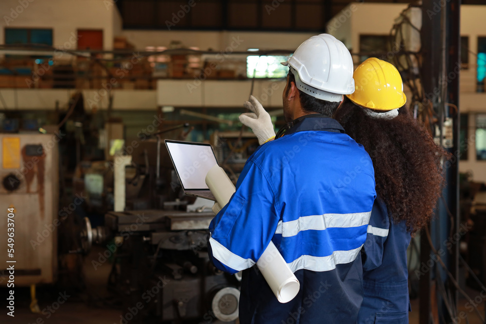 Rear view architect or worker checking at heavy industry manufacturing factory with walkie talkie radio and blueprint. Multiethnic business manager looking in future with warehouse factory building