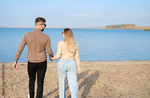 A young couple in jeans and sweaters hold hands and walk along the beach. Back view. Sunny weather
