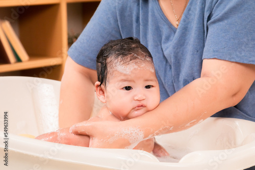 Asian newborn baby in bathtub taking a shower filled with soap bubbles, single mother bathing daughter and washing baby head with warm water with carefully and love, hygiene concept.