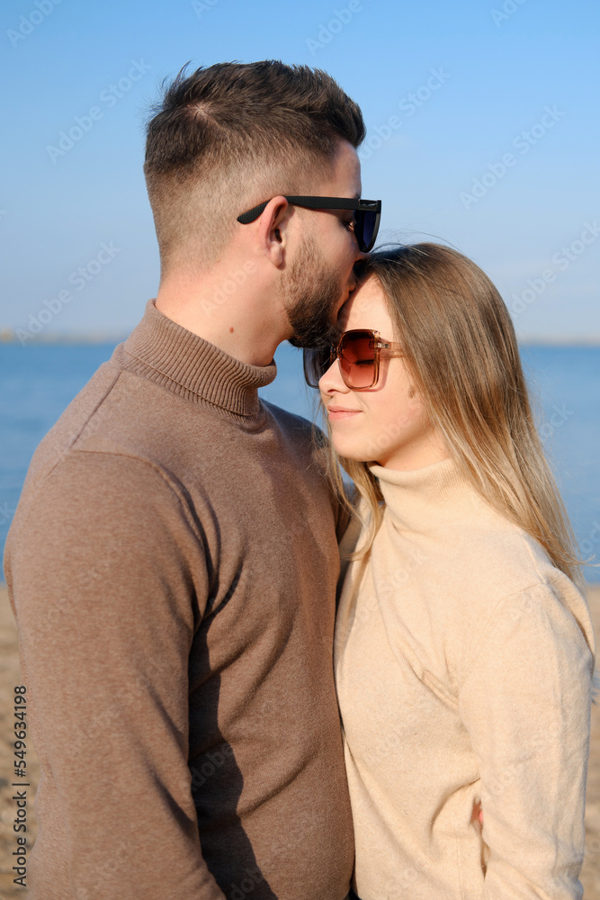 Beautiful young couple posing against the blue sky and water. The guy hugs the girl and kisses on the forehead Beautiful young couple posing against the blue sky and water. The guy hugs the girl and kisses on the forehead