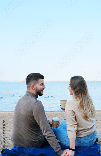A young man and woman are sitting on the beach with their backs to the camera with glasses of coffee in their hands. Date by the lake