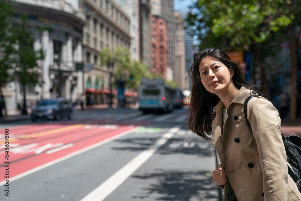 smiling asian chinese woman commuter looking into space while waiting for bus at roadside on a sunny day in san Francisco California usa