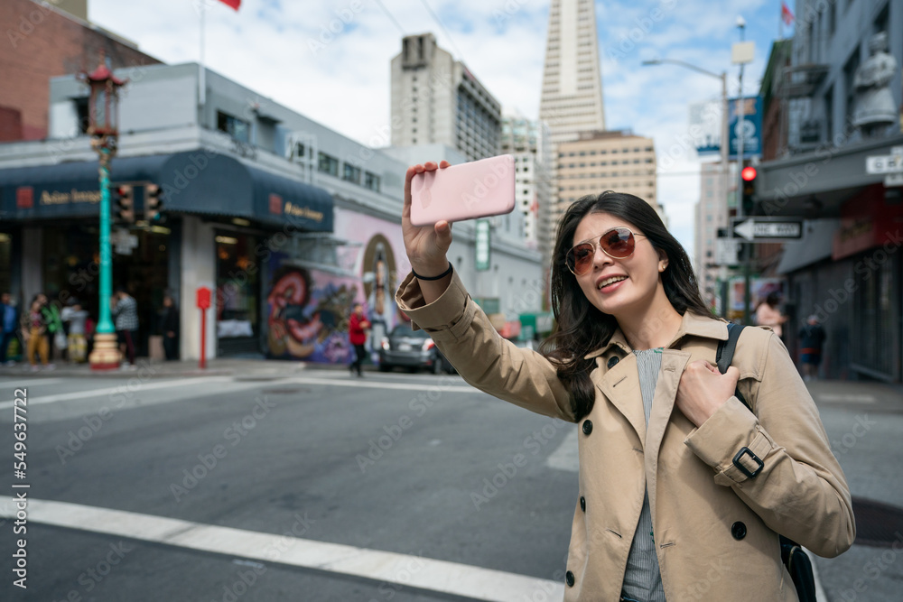 cheerful asian chinese girl tourist wearing sunglasses having video call with friend on cellphone while traveling in financial district of san Francisco's Chinatown in California usa