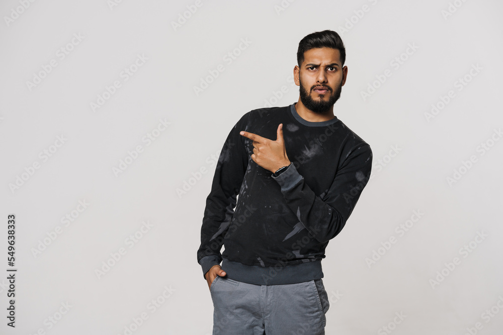 Young indian man with beard frowning and pointing finger aside Young indian man with beard frowning and pointing finger aside