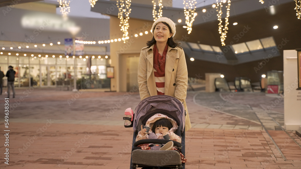 cheerful asian mother taking evening walk with her baby daughter in the pushchair outside a shopping center with Christmas light during the winter holiday