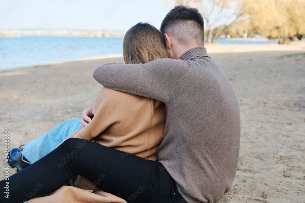 Fototapeta premium The guy hugs the girl by the shoulders. The couple sits with their backs to the camera. Date on the beach in the cold season