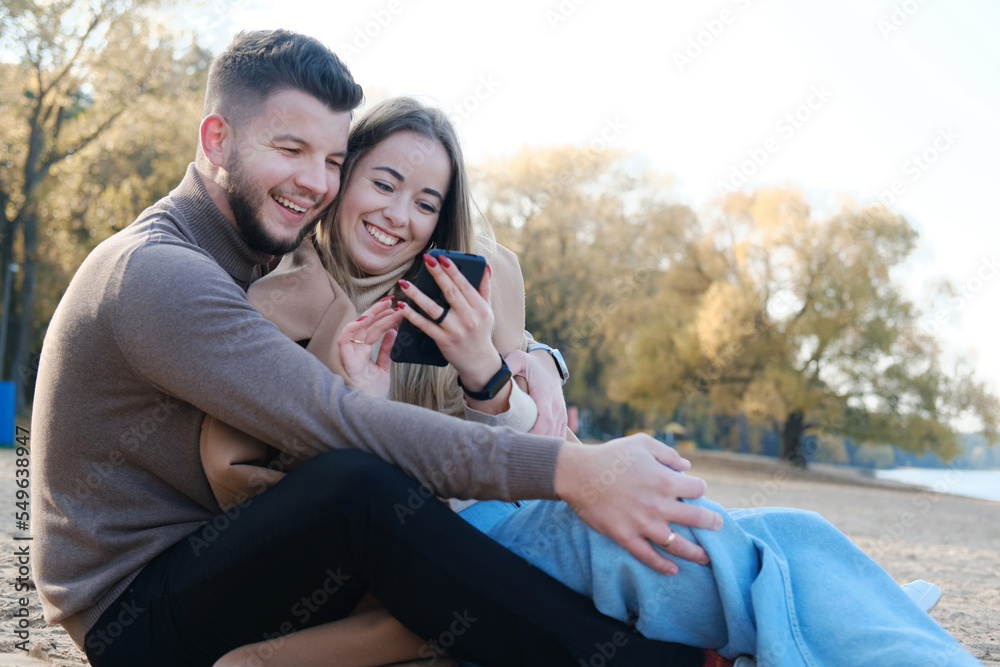 A guy and a girl are sitting on the beach, looking at the phone and laughing A guy and a girl are sitting on the beach, looking at the phone and laughing