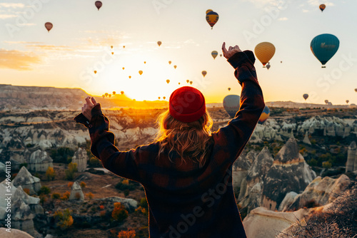 A girl in a red hat and a plaid shirt stands with her back against the background of dawn and hot flying balloons