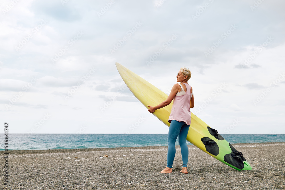 Young blond woman surfer with surfboard on the beach in cloudy day. Extreme sport, water sports concept.