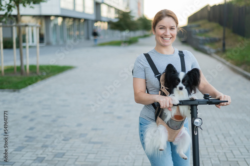 A woman rides an electric scooter with a dog in a backpack. Pappilion Spaniel Continental in a sling. photo