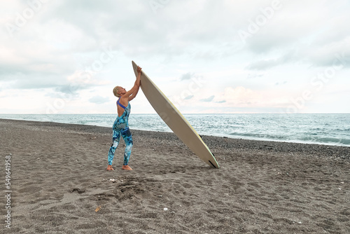 Young blond woman surfer with surfboard on the beach in cloudy day. Extreme sport, water sports concept.