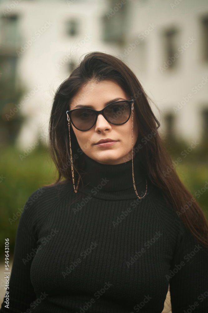 Fashionable and sexy portrait of a brunette woman. The confident and trendy young girl wears sunglasses and chains. Woman with long natural dark hair.