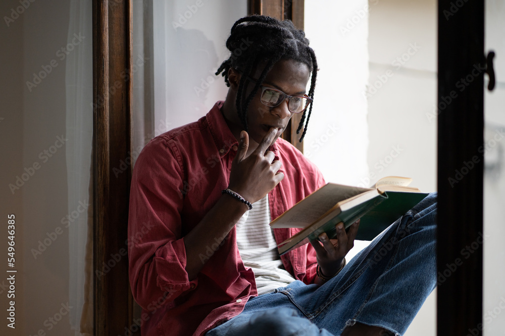Young focused black man wearing eyeglasses learning foreign language by reading books, African American student guy sitting on windowsill at home Learning from textbook. College exams preparation - obrazy, fototapety, plakaty 