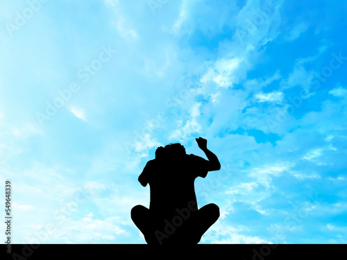地面に座って腕を振り上げている男性シルエット_青空背景