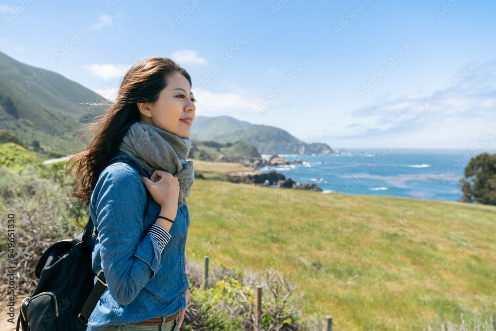 smiling asian Taiwanese female traveler with backpack looking at the view of sea on grassland on a windy day in big sur California usa