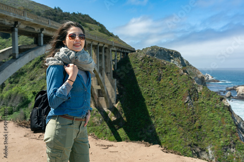 stylish asian Japanese female traveler enjoying cool breeze and warm California sun at seaside cliff with big sur bridge at background