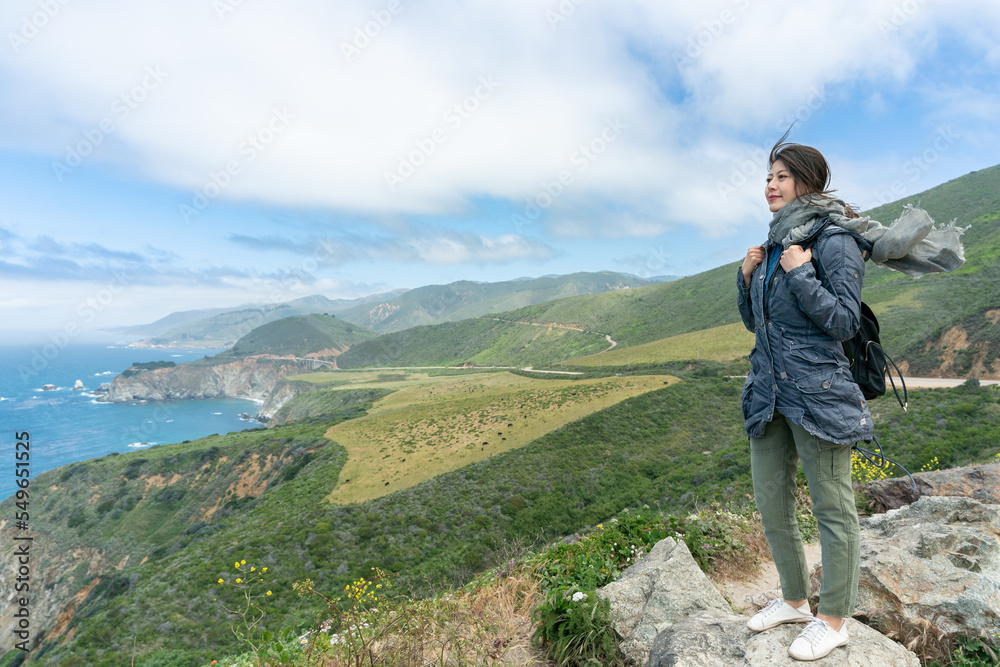 full length of smiling asian Japanese woman traveler on adventurous hiking trip in big sur. sheâs enjoying natural beauty of pacific coastline on top of hill in the wind