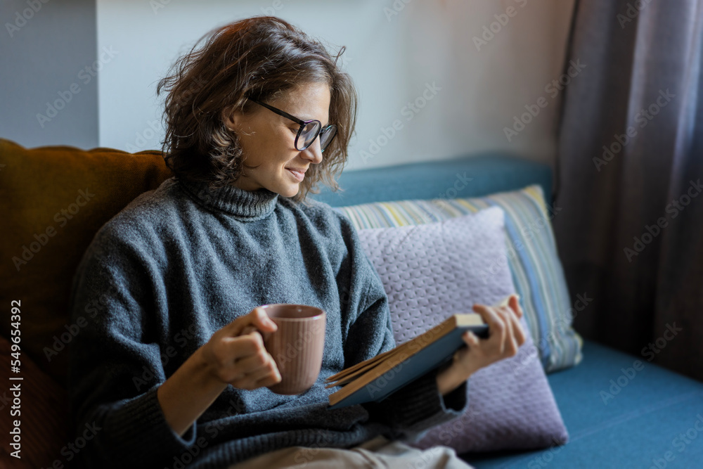 Young smiling cheerful woman in a warm sweater and eyeglasses reading a book while sitting on the sofa with a mug of coffee