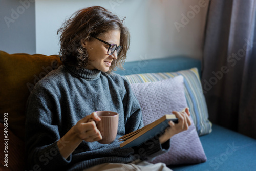 Obraz na płótnie Young smiling cheerful woman in a warm sweater and eyeglasses reading a book while sitting on the sofa with a mug of coffee w salonie