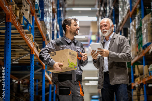 Young wearhouse worker walking accross wearhouse streets with his senior manager, discussing about problems in company and prepairing boxes for delivering