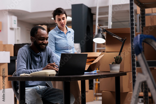 Fashion warehouse manager putting trendy blouse in carton box while supervisor man analyzing shipment on laptop computer before delivering order. Team working at transportation logistics in storehouse