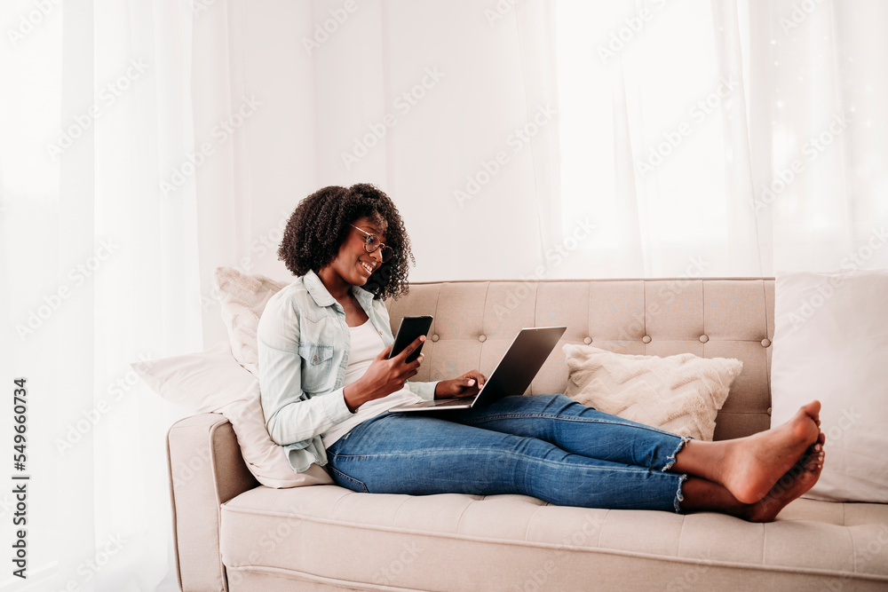 Young freelancer with smart phone and laptop sitting on sofa at home