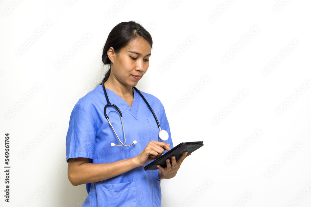 Woman physical therapist in blue uniform and stethoscope typing on tablet computer. Stand in front of white wall. Woman physical therapist in blue uniform and stethoscope typing on tablet computer. Stand in front of white wall.