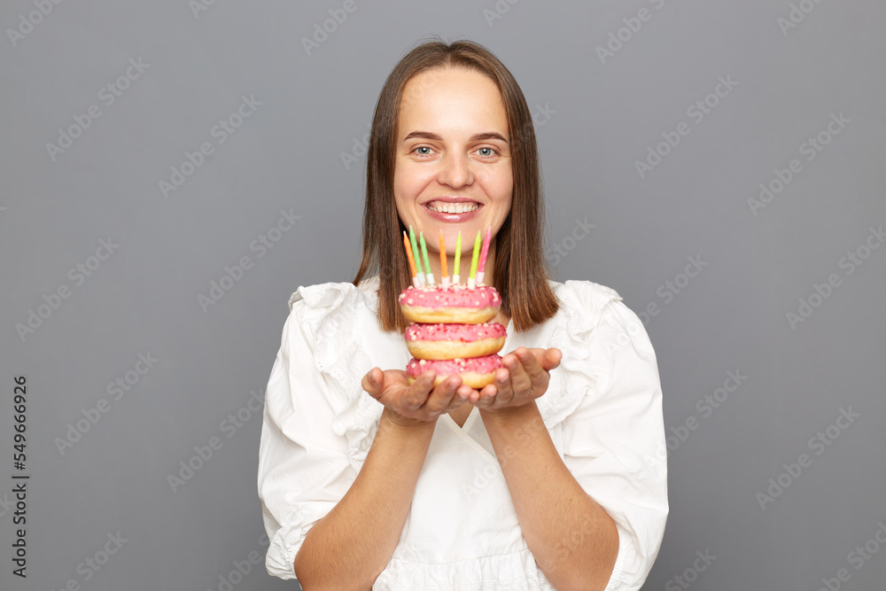 Indoor shot of pretty friendly pleased woman with brown hair wearing white blouse, looking at camera with happiness, holding donut with candles isolated over gray background. Indoor shot of pretty friendly pleased woman with brown hair wearing white blouse, looking at camera with happiness, holding donut with candles isolated over gray background.