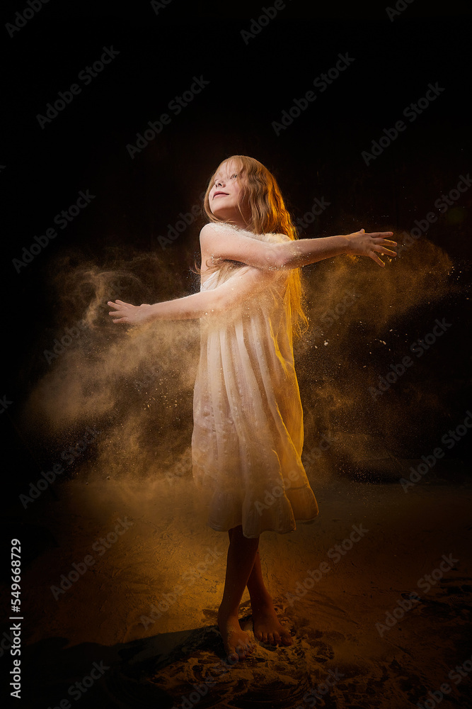 Fototapeta premium A girl in a white shirt and with long hair, looking like a ghost. A young model at a photo shoot with smoke, fog, flour, dust. A little fairy girl on a dark black background