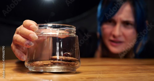 Woman found large cockroach in glass jar of sweet cookies, looking at insect with disgust. Selective focus on foreground at transparent container, in background blurred face of housewife photo