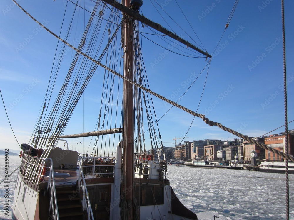 Obraz premium Aker Brygge in Oslo with a sailing ship lying in the Oslofjord with ice floes, Norway, on a beautiful cold sunny day with blue sky.