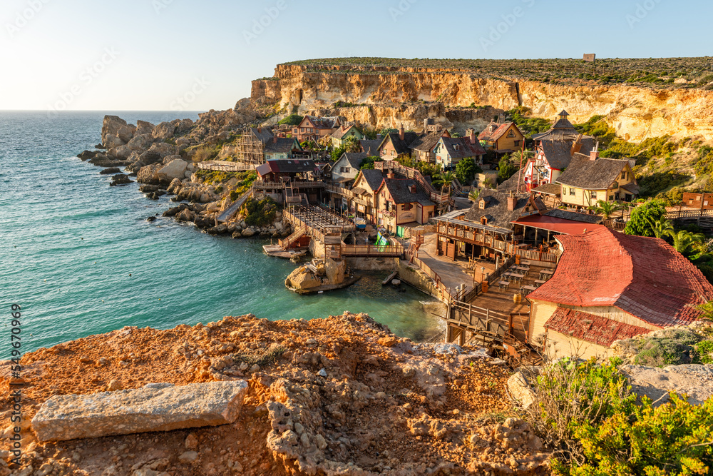 Fototapeta premium Malta, Il-Mellieha. View of the famous Popeye Village in Anchor Bay. Turquoise sea waters, blue sky and sunset light on cliffs