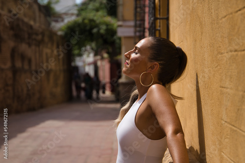 Young and beautiful woman, with ponytail, wearing a white top, eyes closed, leaning against a wall, relaxed, receiving the sun rays. Concept beauty, sun, relax, peace, tranquility. © Manuel