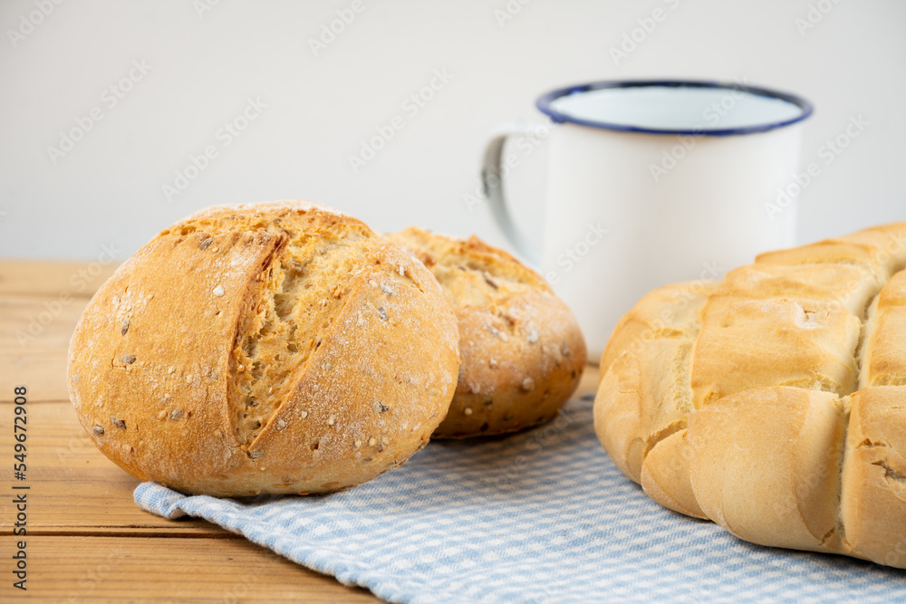 Close-up of loaf of rustic bread and rolls on cloth and wooden table with knife and cup, white background, horizontal, with copy space