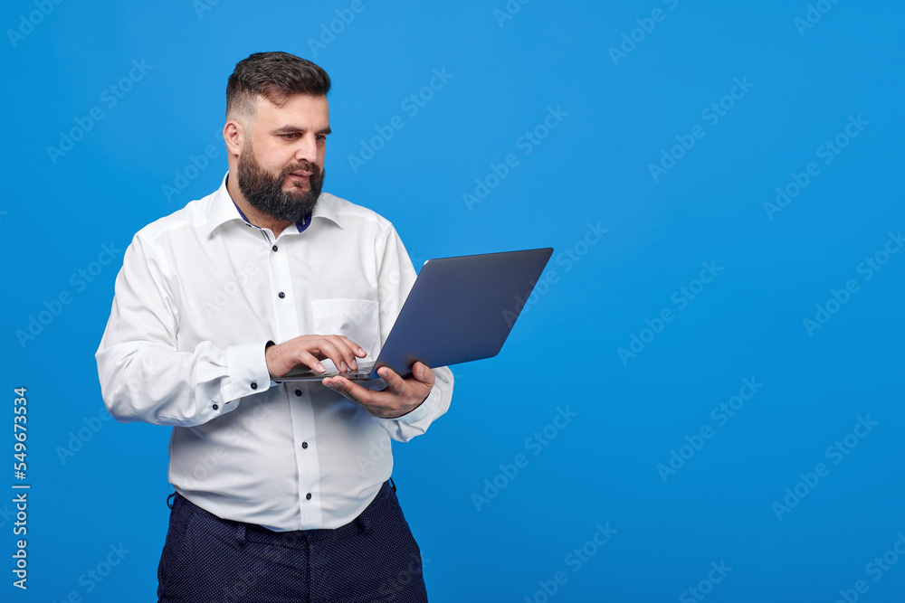 A bearded man holds a laptop in his hands on a blue background. A solid man in a white shirt works at a laptop and looks into the camera.
