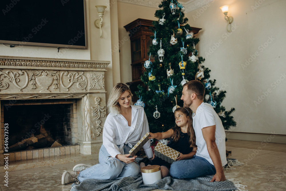 Obraz premium A daughter opens a gift next to her parents on the background of a Christmas tree