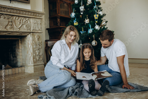 A daughter opens a gift next to her parents on the background of a Christmas tree © Roman