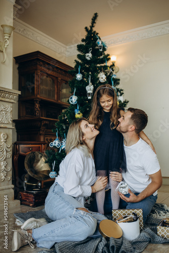 A daughter opens a gift next to her parents on the background of a Christmas tree © Roman