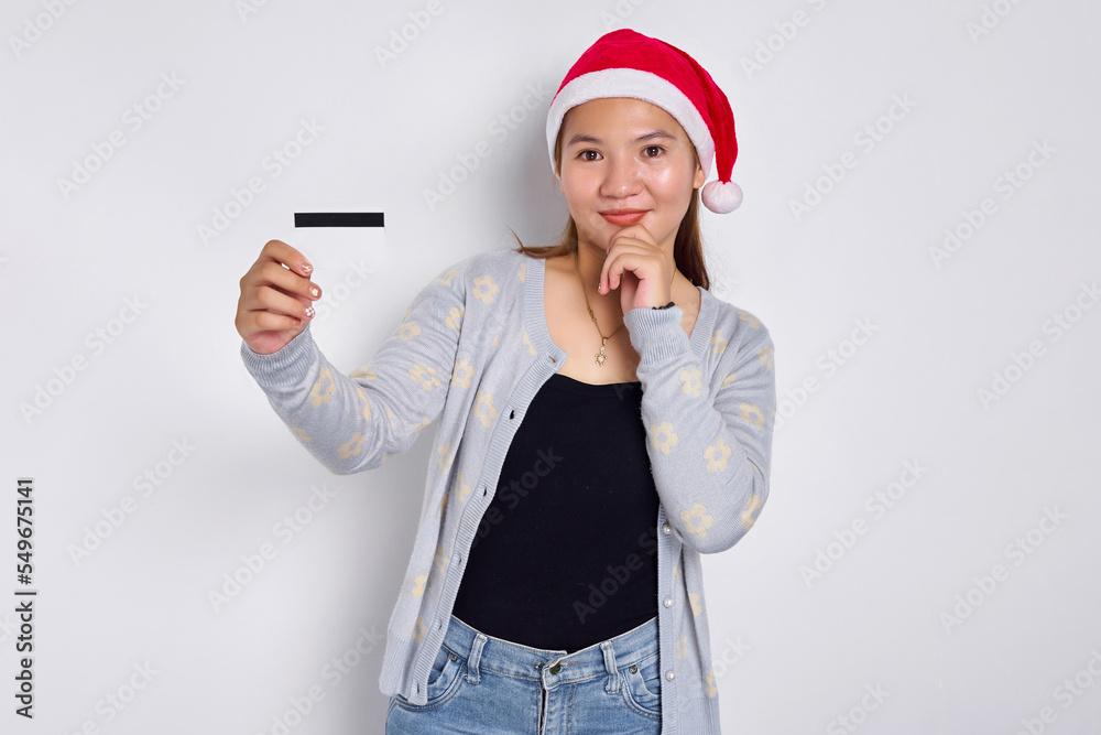Pensive young Asian woman in a Christmas hat showing blank white credit card and touching chin on hand isolated on white background Pensive young Asian woman in a Christmas hat showing blank white credit card and touching chin on hand isolated on white background