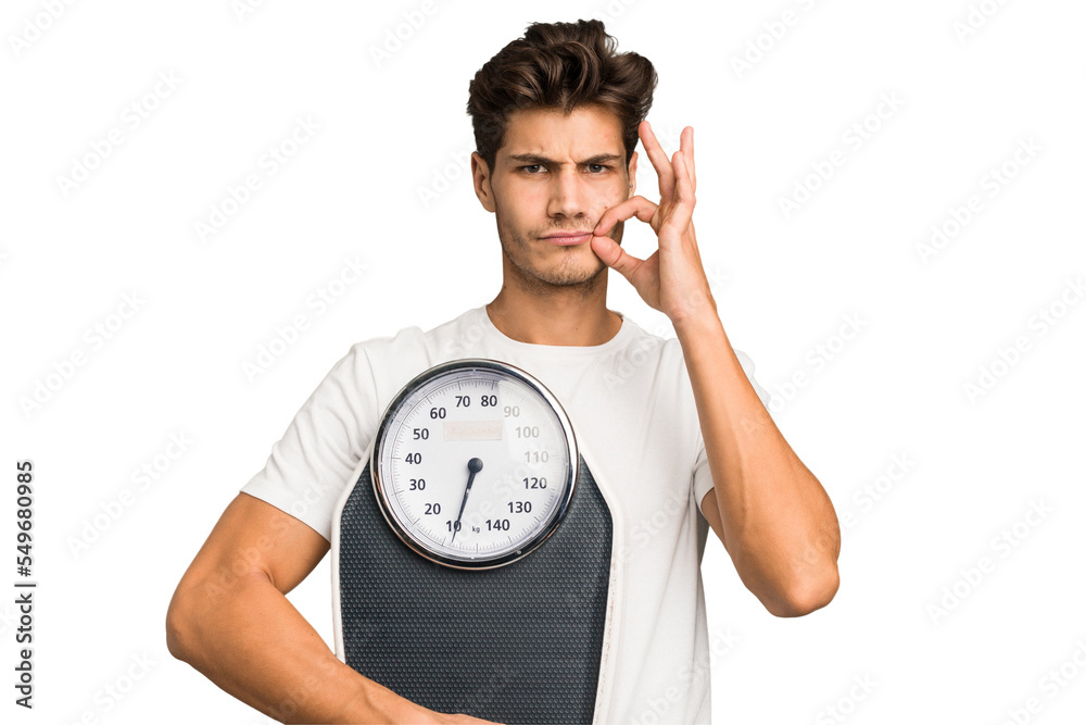 Young caucasian man holding a scale isolated with fingers on lips keeping a secret. Young caucasian man holding a scale isolated with fingers on lips keeping a secret.