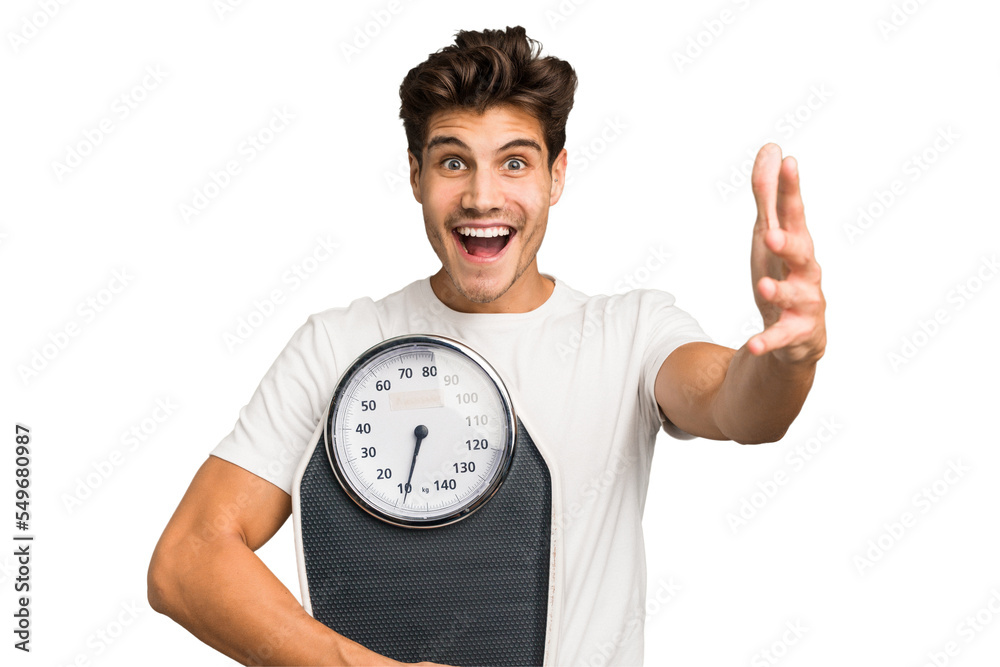 Young caucasian man holding a scale isolated receiving a pleasant surprise, excited and raising hands. Young caucasian man holding a scale isolated receiving a pleasant surprise, excited and raising hands.