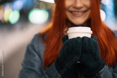 Girl drinking hot coffee while walking in the evening city market decorated with holiday lights in the evening. Feeling happy in big city. Spending winter vacations