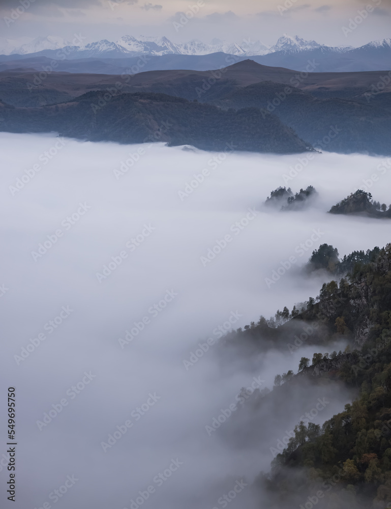 Low clouds in the mountains filled the entire valley and rocky peaks and hills with trees stick out above the clouds, mountains with snow in the background, fog between the mountains on an autumn