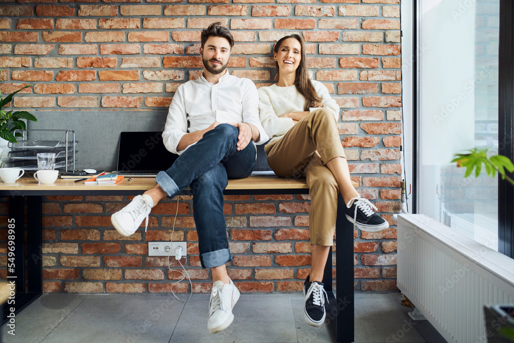 Obraz premium Smiling young entrepreneurs business partners sitting together on the desk in small startup office