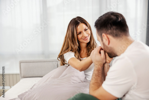 A man in pajamas is kissing his woman's hand while sitting in their bed in bedroom.