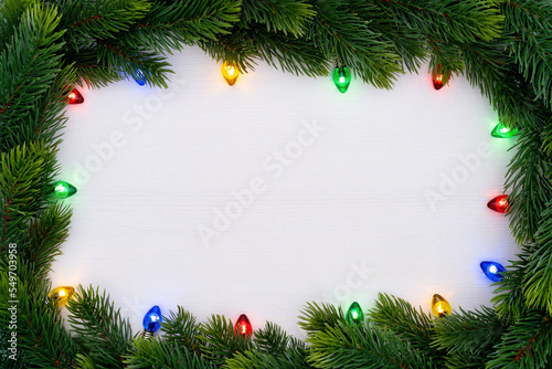 Christmas theme. Frame made of fir branches with glowing retro garland. Nostalgic multicolor illuminated garland on a white wooden background with copy space.
